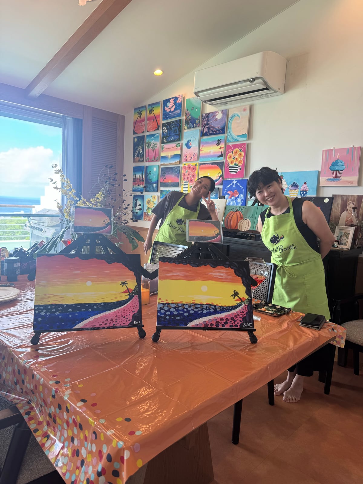 Two Pinto Beetle guests smiling with finished sunset beach paintings in the ocean-view studio